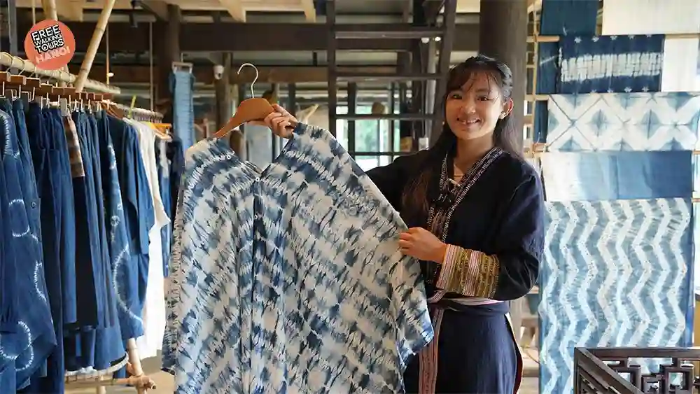 Smiling Hmong artisan holding up a handmade indigo tie-dye shirt inside a Sapa craft shop filled with batik fabrics and indigo garments