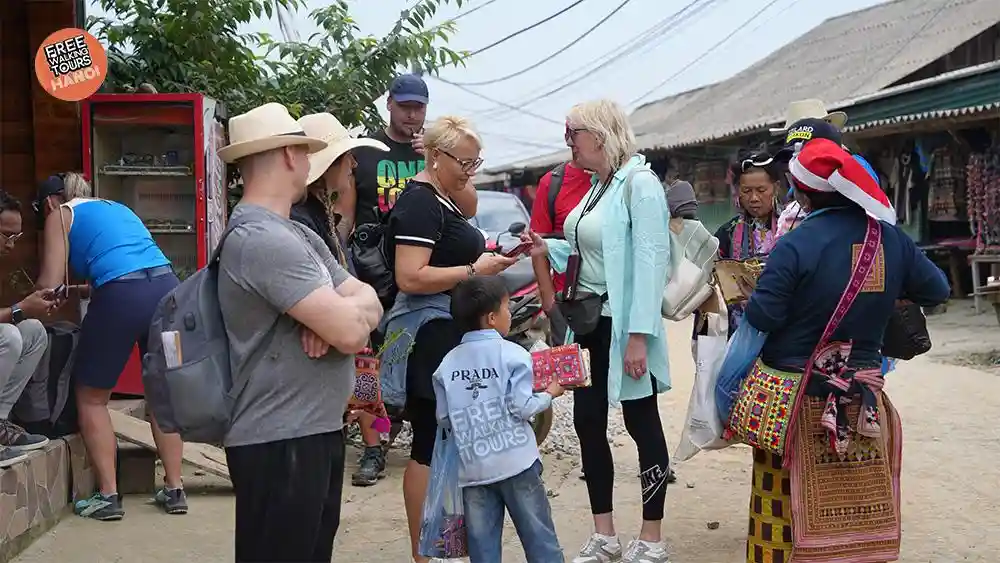 International travelers gathering with Hmong vendors and a small child on a street in a Sapa ethnic village before starting a free walking tour
