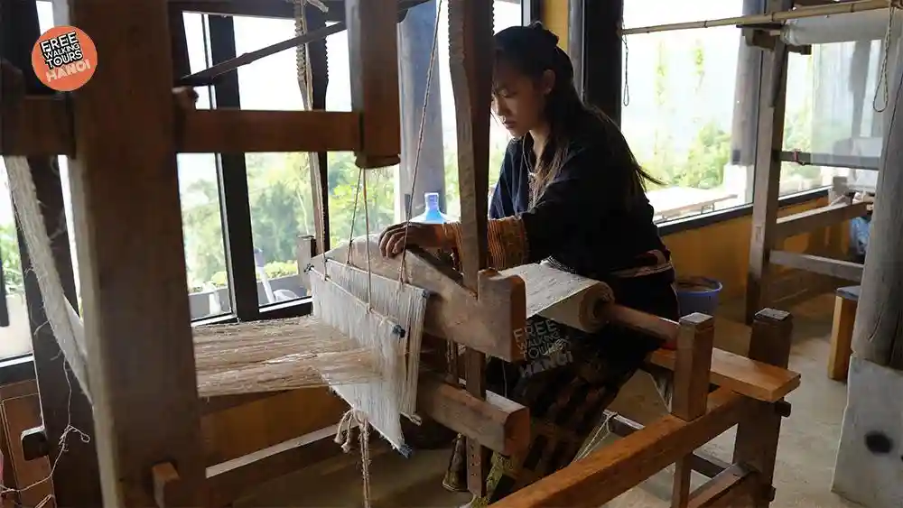 Young Hmong woman weaving hemp fabric on a traditional wooden loom inside a Sapa village workshop, with valley views through the windows