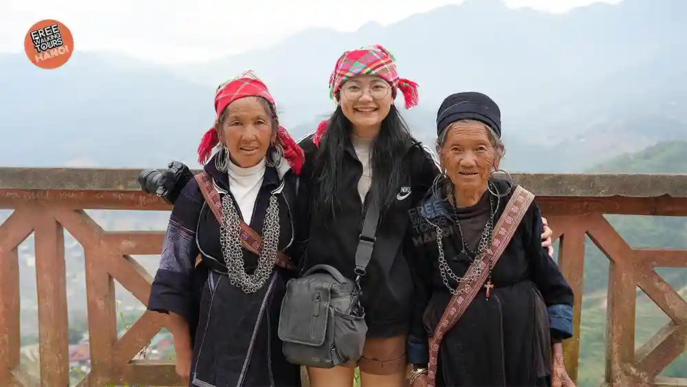 Young traveler standing between two Red Dao women elders in traditional indigo dress and red headscarves at a Sapa valley viewpoint